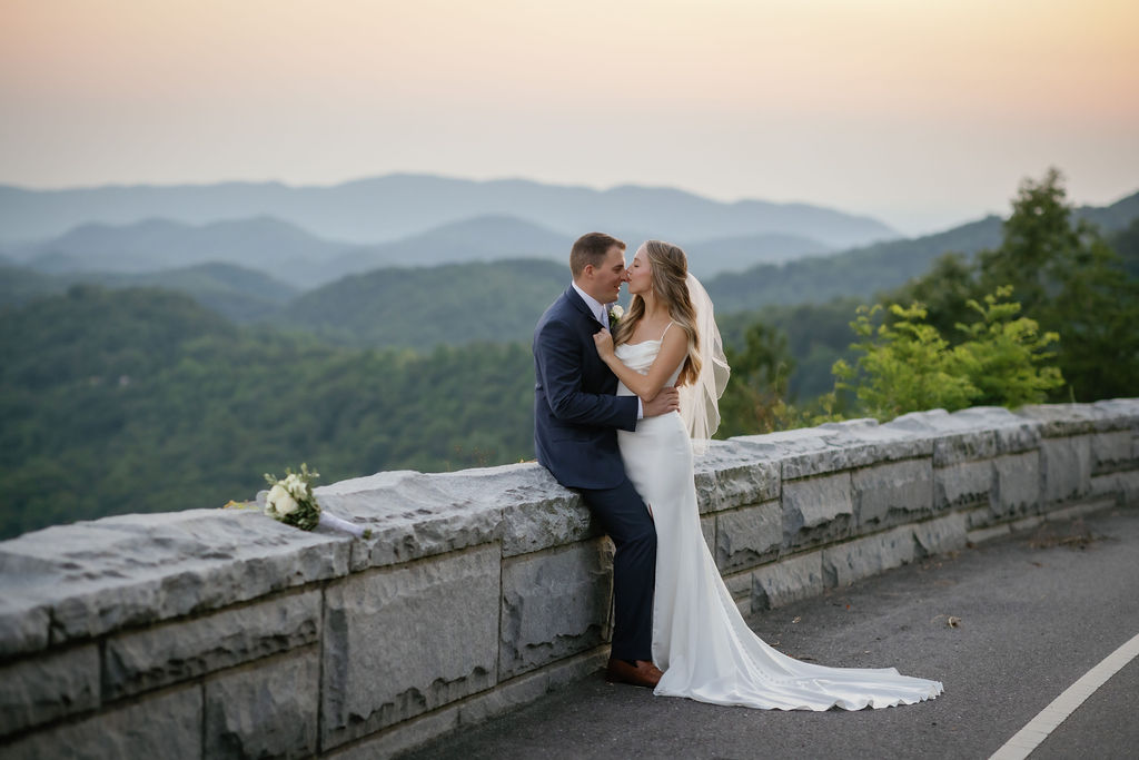 foothills parkway elopement with bride leaning into her bride as he sits on a stone divider at sunset 