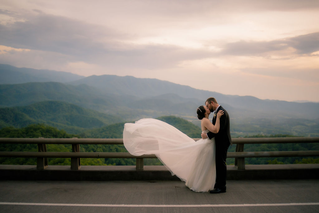 whle eloping to Gatlinburg bride and groom kiss passionately at the foothills parkway with the Smoky Mountains standing tall behind the couple right after sunset