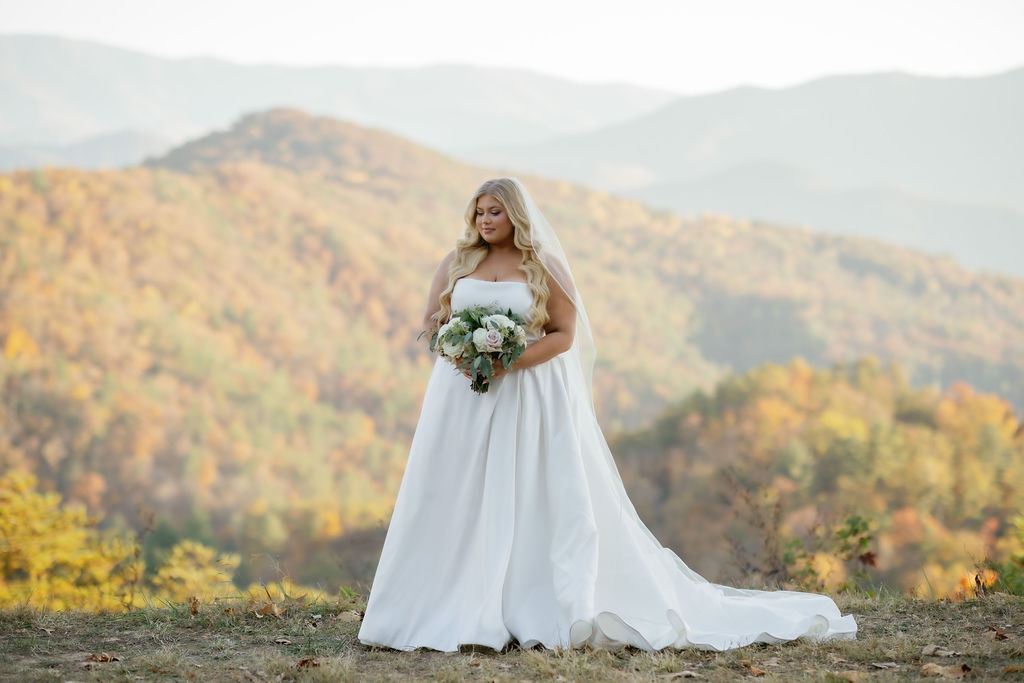 fall elopement in Gatlinburg bridal portraitn with plus size bride in a satin gown posing on an outlook in the foothills with rolling hills of autumn colored trees behind her captured by photographers in gatlinburg tn